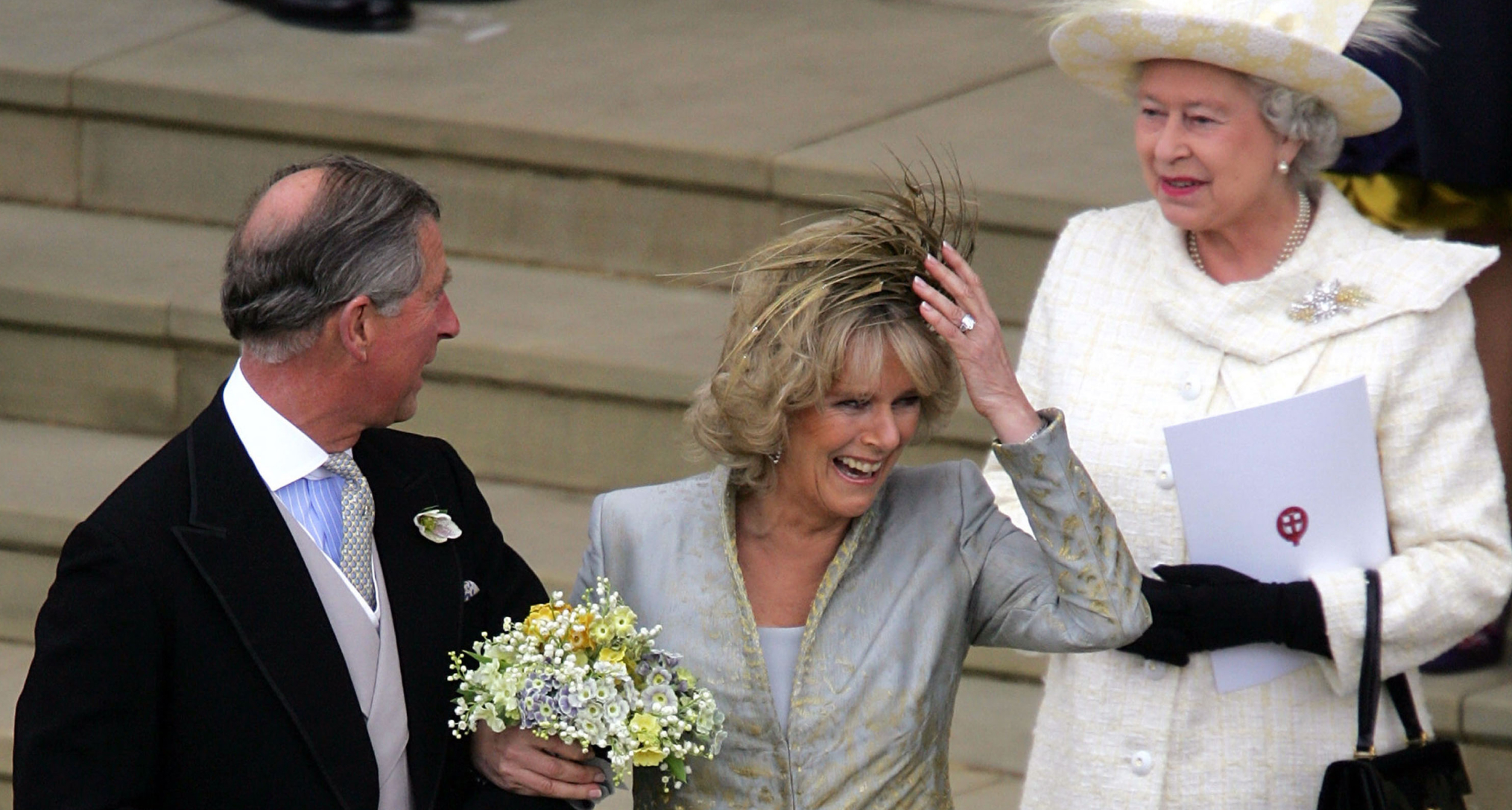 King Charles linking arms with Queen Camilla, holding on to her hat on her wedding day, with Queen Elizabeth standing behind her