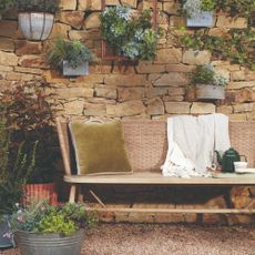 A garden with a garden bench with a rattan backrest set against a stone wall with hanging planters