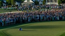 A long-range shot of Rory McIlroy down on his knees and arms on the 18th green at Augusta National with the large crowd cheering in the background