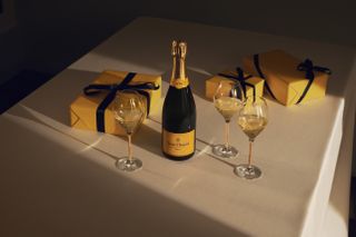A bottle of Veuve Clicquot Yellow Label on a white tablecloth with Champagne glasses and wrapped presents