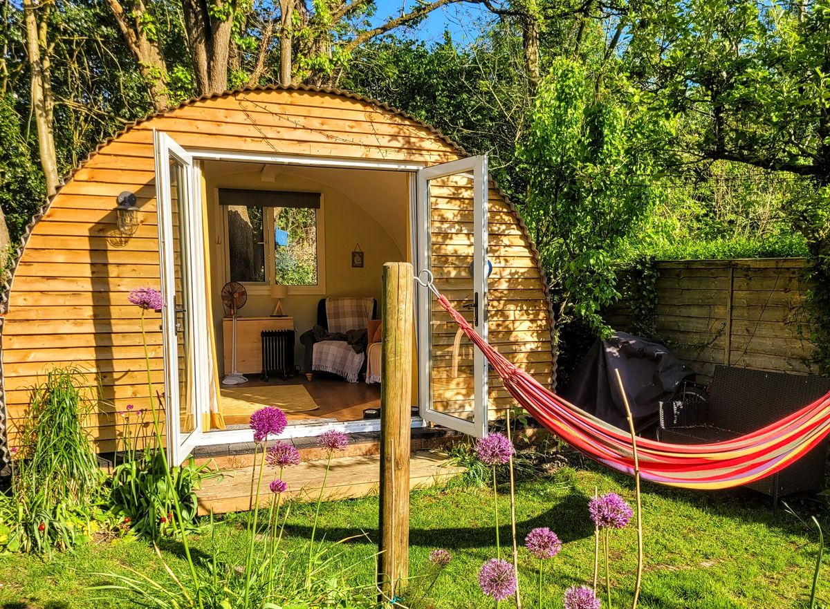 A hammock lies in front of this wooden hanger shelter converted into a shed with mossy plants lying either side