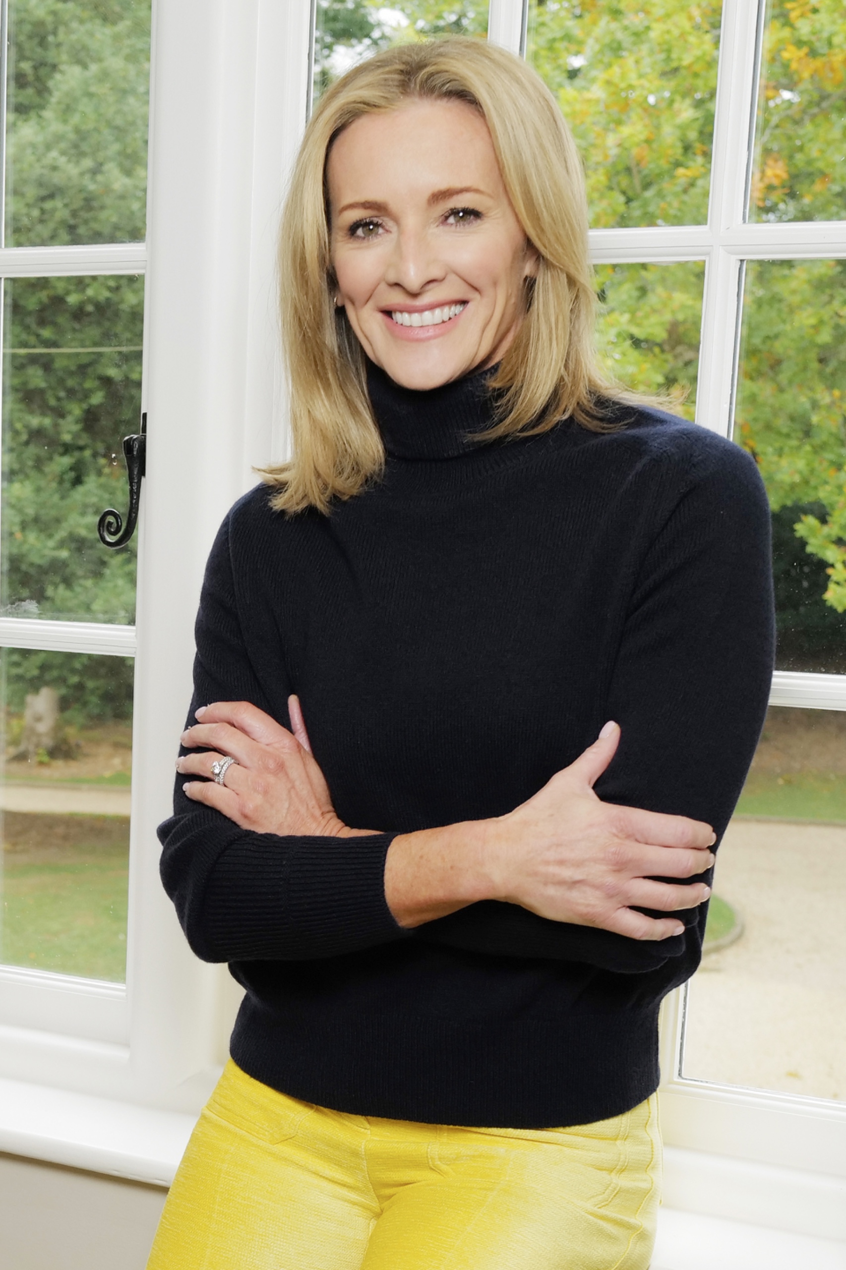 A woman leaning on a windowsill with her arms crossed, smiling at the camera and wearing a black jumper and yellow trousers
