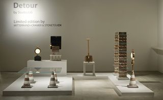 A display of street cones and bricks made out of pebbles on a geometric shaped white platform. A square shaped glass is place on the cones (made from pebbles) to create a table. A stack of 2 bricks on the bricks (made from pebbles) divided by clear glasses.