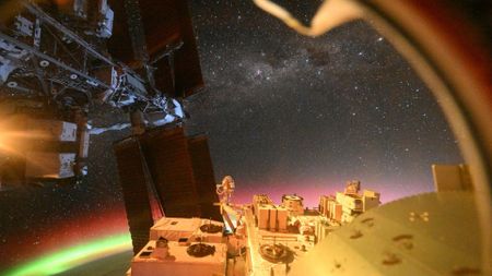 A view from the ISS that shows the ISS solar panels, auroras over Earth, and outer space with a vast array of stars. 