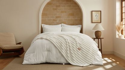 A white Bedsure 2-in-1 comforter on a bed in a white bedroom. Behind the bed is an exposed brick arch, and to the right is a wooden bedside table with a brown lamp with white shade, and above is a framed artwork. To the right is a window, and to the left of the bed is a low, cushioned beige chair with a brown wood frame. The floor is wood, with a beige rug on top.