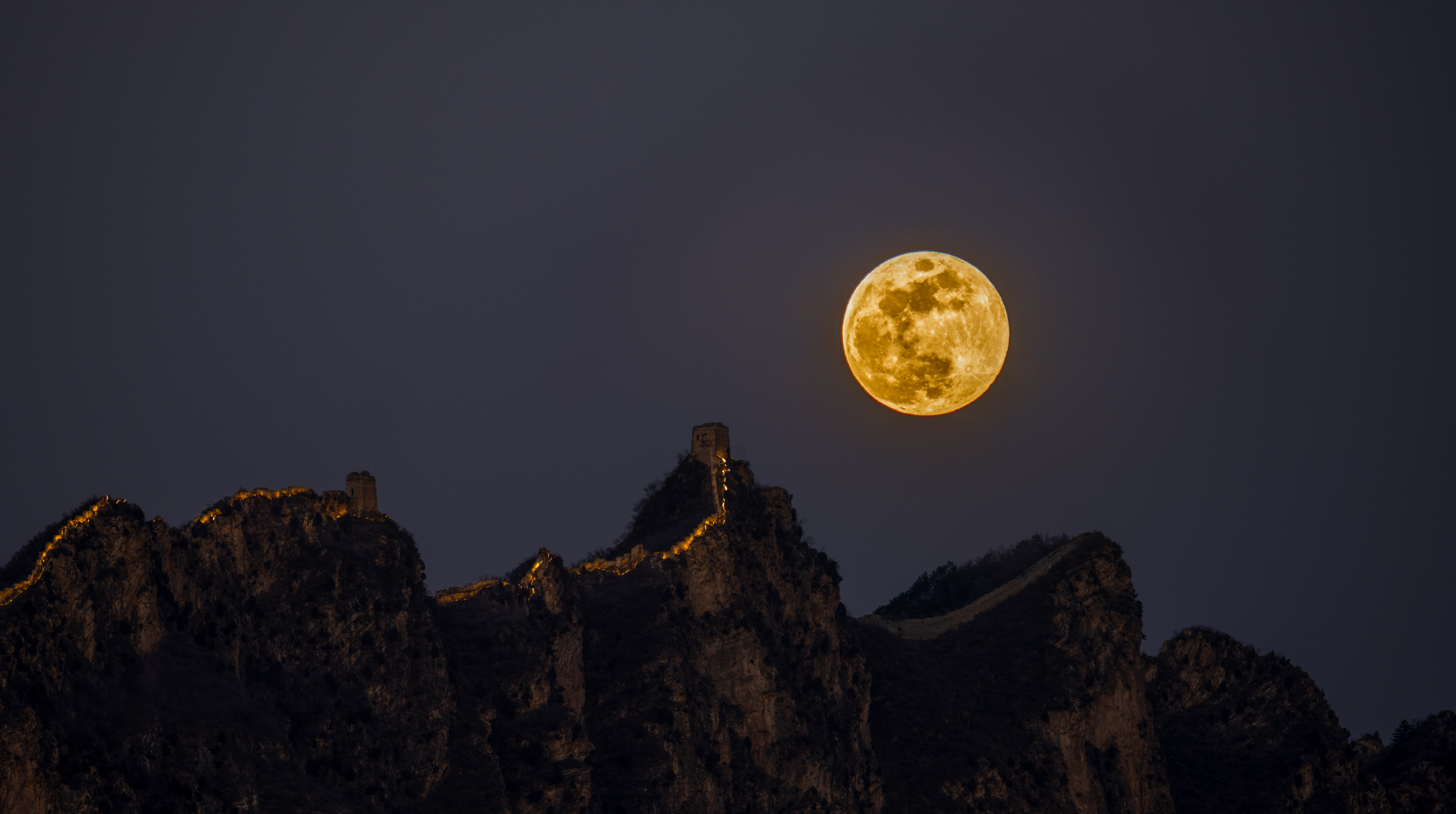 a bright yellow hued full moon shines over the great wall of china snaking it's way over the mountain tops.