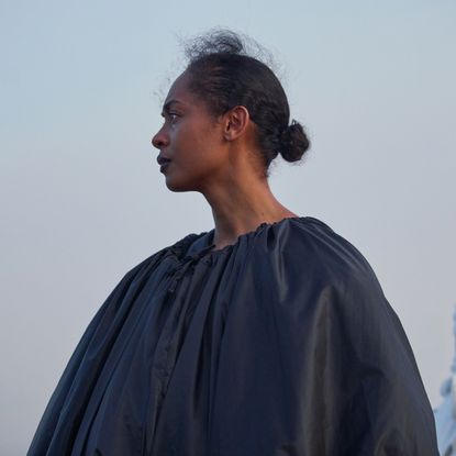 A woman (Susan Chardy as Shula) stands outdoors in the morning, with a man covered in sanitary pads behind her (right), in 'On Becoming a Guinea Fowl.'