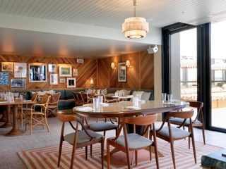 Interior view of the dining area on the 10th floor at White City House featuring grey flooring, an orange and off-white coloured geometric rug, wood panelled walls, wall lights, wooden dining tables with tableware, wooden and wicker chairs, a white ceiling, a pendant light, wall art and lamps, dark coloured corner seating with contrasting cushions and glass doors