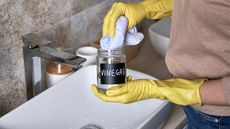 Person with yellow gloves holding a bottle of vinegar over a sink, preparing to clean