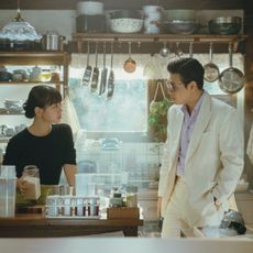 Two people (Go Min-si and Kang Ha-neul) stand in a kitchen with an open back window, in the Korean drama 'Tastefully Yours.'