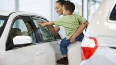 A young woman holding a toddler looks at pricing information on a new car.