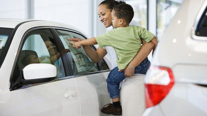 A young woman holding a toddler looks at pricing information on a new car.