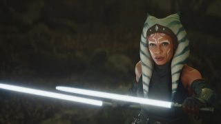 Still from a Star Wars movie. Here we see Ahsoka holding two short white lightsabers. She has orange skin and white and blue striped head tails.