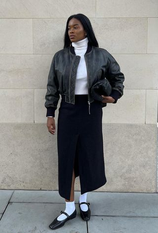 A woman wearing a black leather bomber jacket over a white turtleneck with a denim maxi skirt, white socks, and black flats.