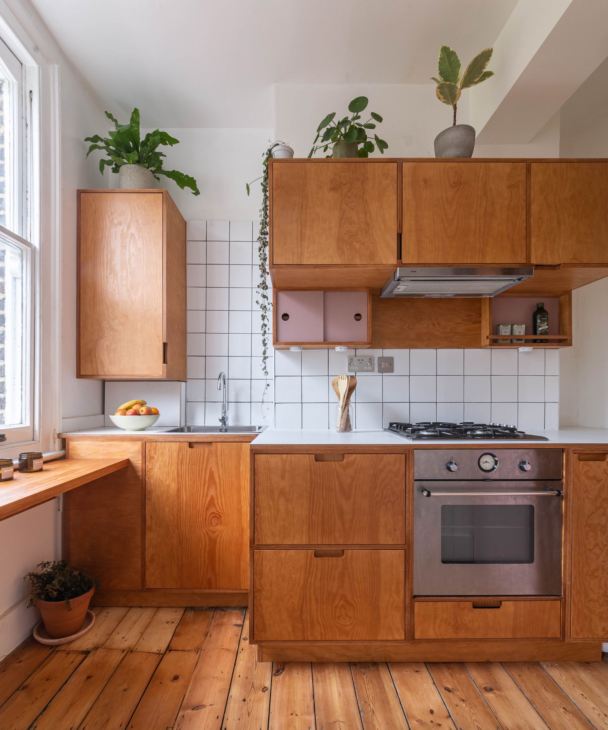 Small, cozy kitchen with plywood cabinetry, white square tile backsplash, rustic wide-plank wood floors, and several potted houseplants.