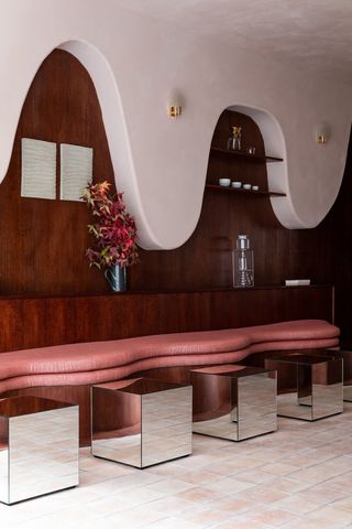 A whimsically designed coffee shop features mirrored chrome cubes as stools, upholstered pink banquettes, wavy textural walls, and wooden surfaces.