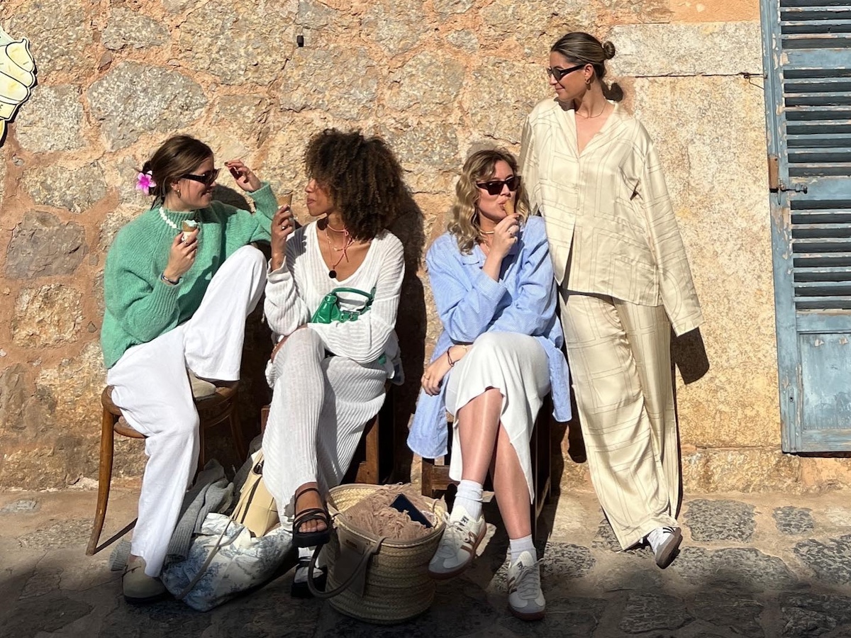 A group of European fashion friends including Amaka Hamelijnck sit on a bench in stylish Scandi-inspired outfits with oversize button-downs, linen pants, and sneakers while chatting and eating ice cream.