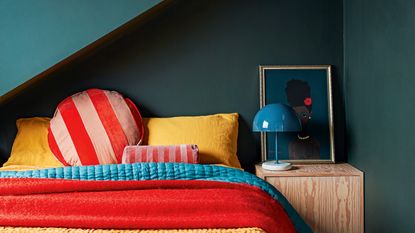 Small bedroom with double bed with black bedhead, pink, green and grey bedding, grey painted floorboards with a rug and retro suitcases at the foot of the bed.