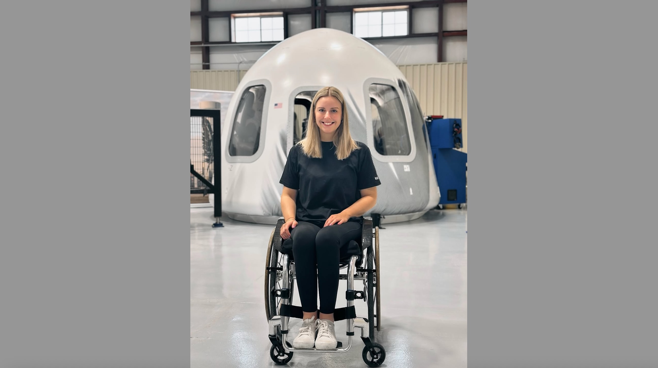 a young woman in a wheelchair sits in front of a white space capsule inside a large hangar