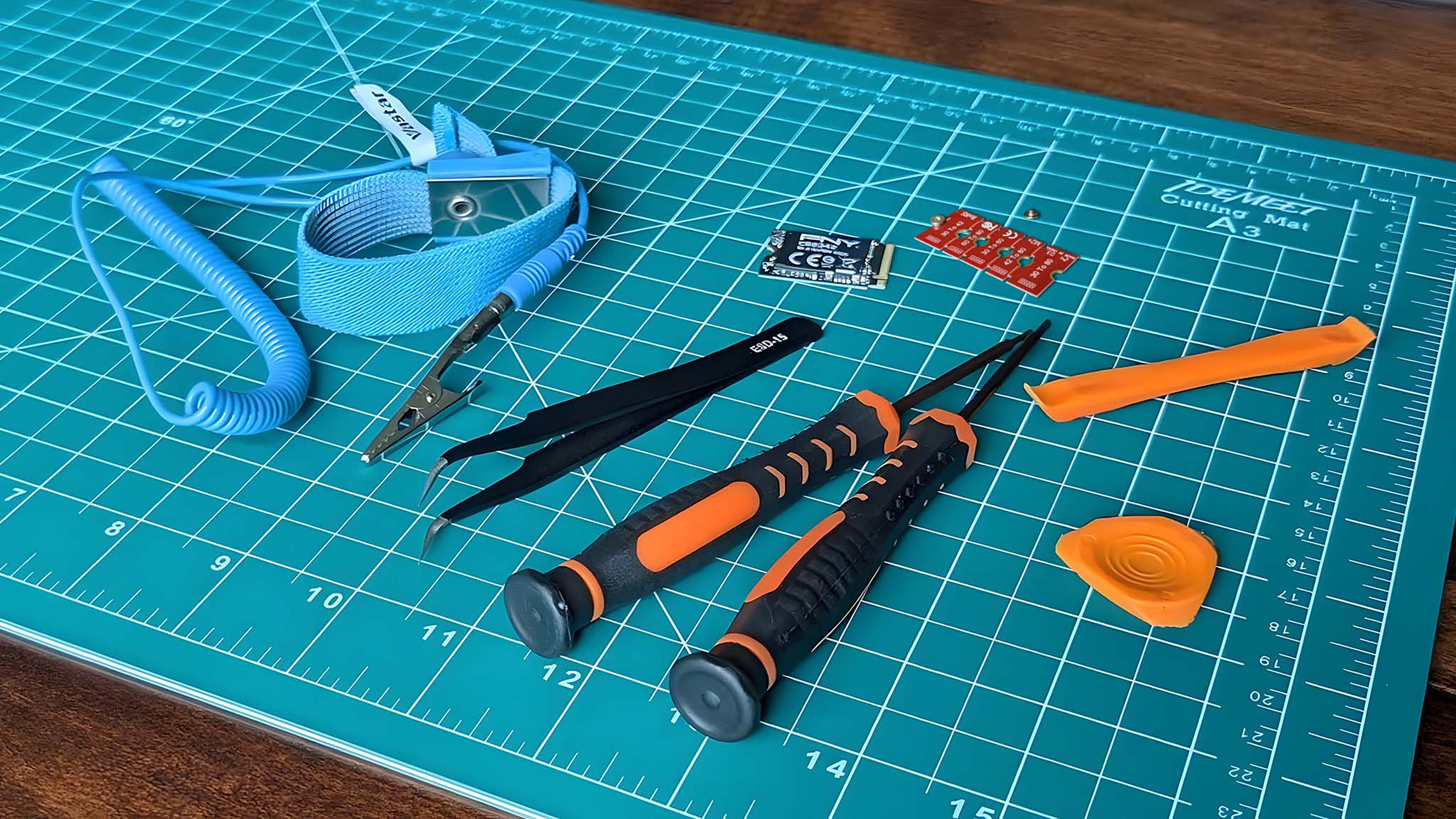 Tweezers, screwdrivers, prying tools, an SSD, SSD extenders, and anti-shock band on a green mat.