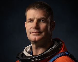 astronaut jeremy hansen looking at the camera. he wears an orange spacesuit and the collar is white. behind him is a black background