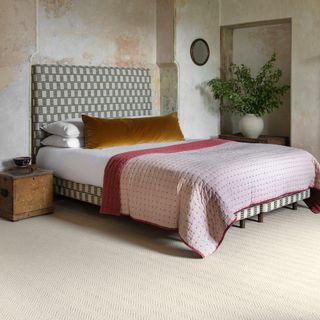 carpet colour trends for 2023, bedroom with cream textured wood carpet, upholstered headboard on bed with green retro fabric, white bedding, mustard velvet cushion, pink quilt, vintage side table, distressed walls
