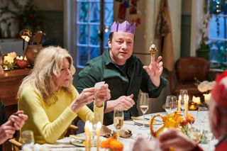 Jamie Oliver sits at a dining table surrounded by guests; the woman next to him is reading a joke from a Christmas cracker. Jamie has a paper party hat on, and is holding up a tiger finger puppet on his index finger