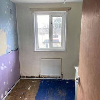 a room with stripped back walls showing old paint and stripped back floors showing floorboards half with blue paint, and a radiator underneath white window showing the outdoors