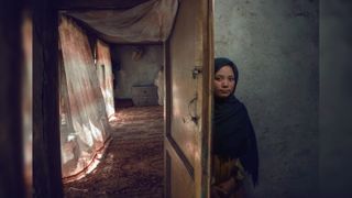 A young girl in a dark headscarf peers out from behind a half-open wooden door with a metal bolt, standing against a dim grey wall, while beyond the door a sunlit room with patterned curtains