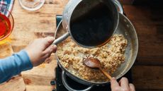 Birds-eye photo of hand pouring stock from a pan into a pan with risotto rice in it. The other hand is stirring the rice using a wooden spoon