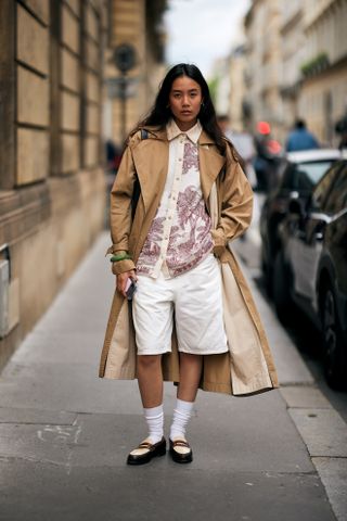 A woman wearing a tan trench coat, red and white button down shirt, white shorts, and loafers.