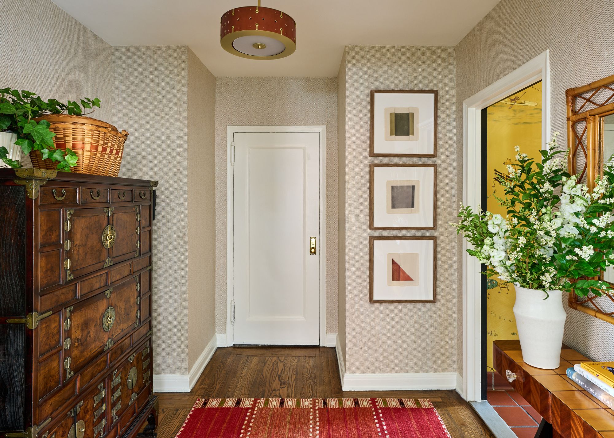 An apartment entryway with neutral walls, wood flooring, and white trim. Framed artworks on the wall, a red rug, and a large, dark antique dresser.