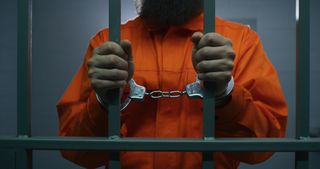Twisted Cover Up airs Saturday, May 25 on Investigation Discovery. Pictured: Criminal in orange uniform and handcuffs holds metal bars, stands in prison cell,