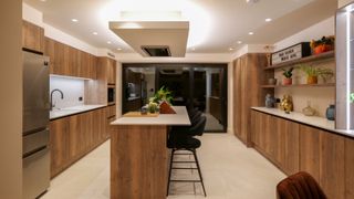 large gallery kitchen with modern wooden cabinetry, central island with ceiling extractor, lighting, cream floor and large stainless steel american style fridge freezer