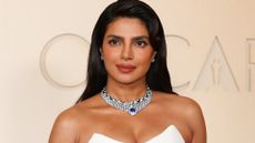 Image of Priyanka Chopra at the 2026 Oscars, smiling at the camera with side-part long brunette hair and wearing a strapless white dress 