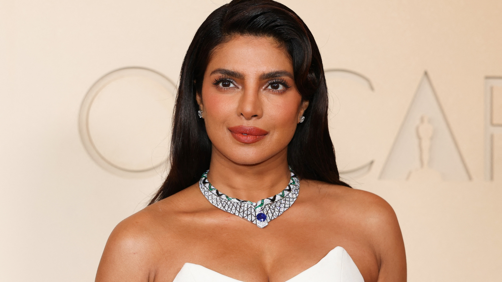 Image of Priyanka Chopra at the 2026 Oscars, smiling at the camera with side-part long brunette hair and wearing a strapless white dress 