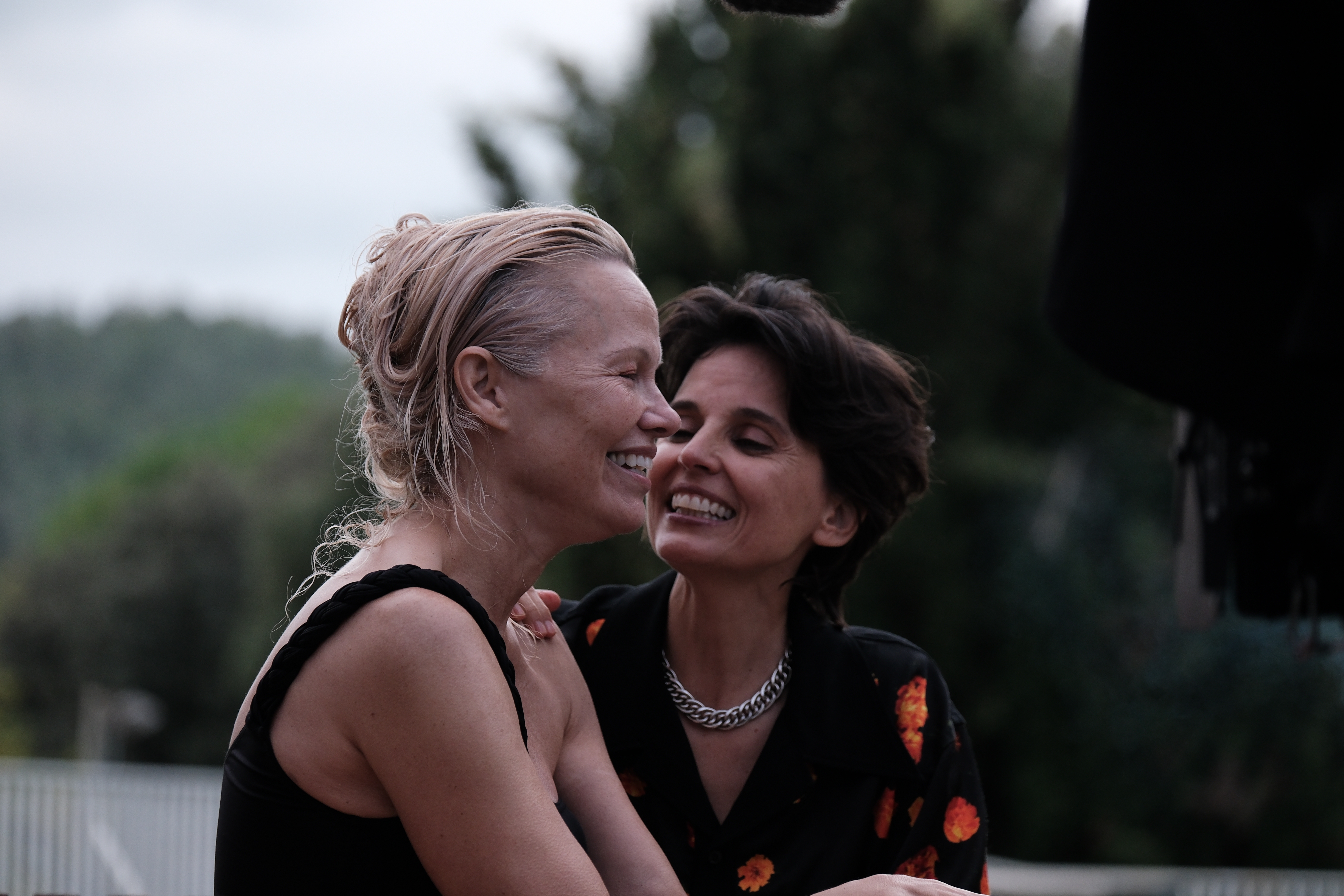 Two women, one blonde in a black swimmingsuit, one brunette with a black shirt and orange patterns and a silver necklace, laugh outdoors.