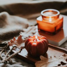 A burning candle in the shape of a tomato, in front of a burning candle in a square jar
