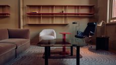 A cool living room with a patterned area rug, modular wall shelving, a glass coffee table, brown couch, white sofa seater, red side table, floor lamps and a black chair