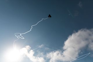 Daytime image, white squiggly digital line attached to a grey inflated aeroglyphic, representing the trail when in flight, blue sky, fluffy white clouds, bright white sun glow bottom left of the shot