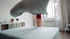 A woman stands next to a mattress in a bedroom, fluffing out a sheet to place on top of the bed