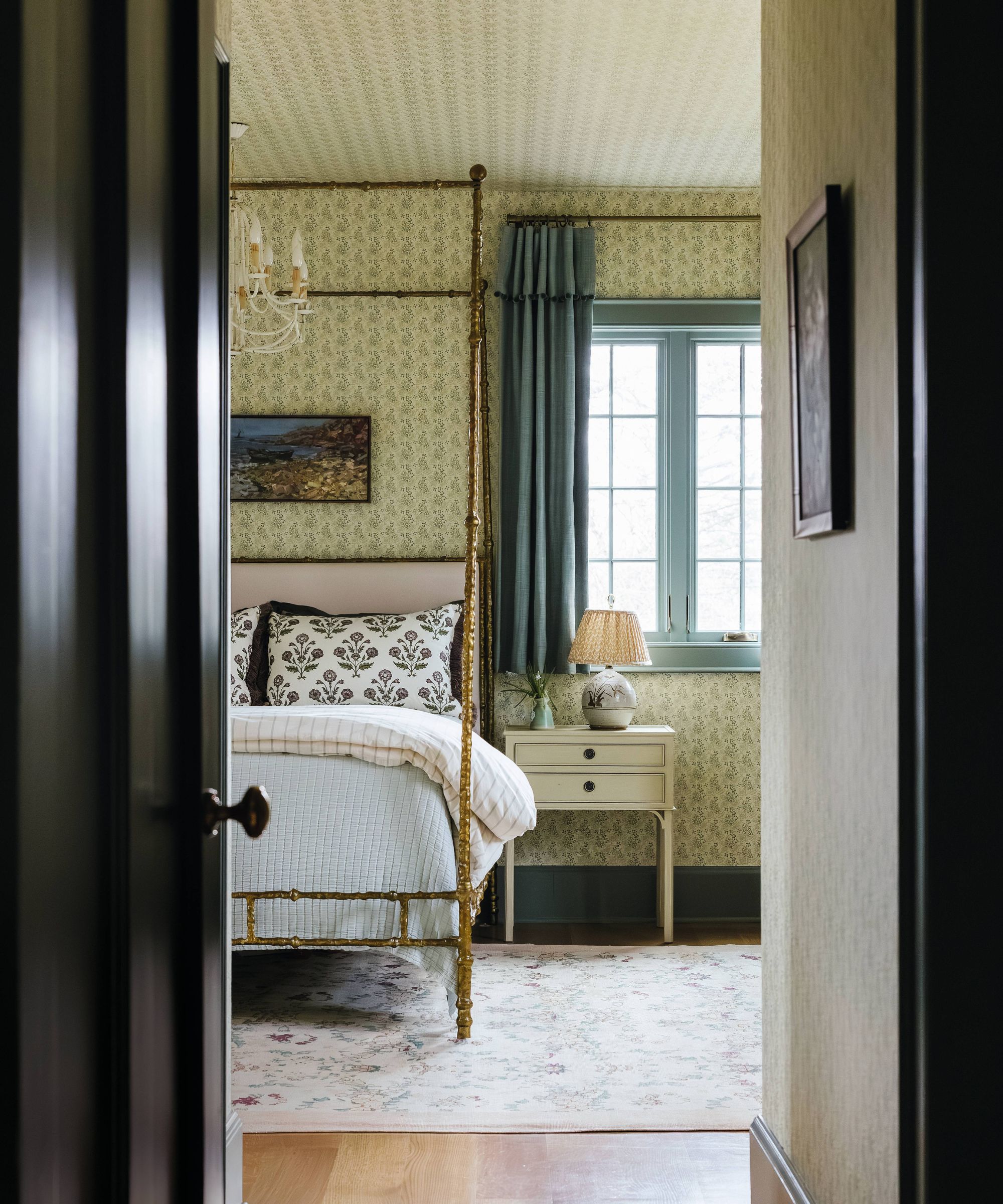 a ditsy floral wallpapered bedroom with brass four poster bed and light trim and drapes