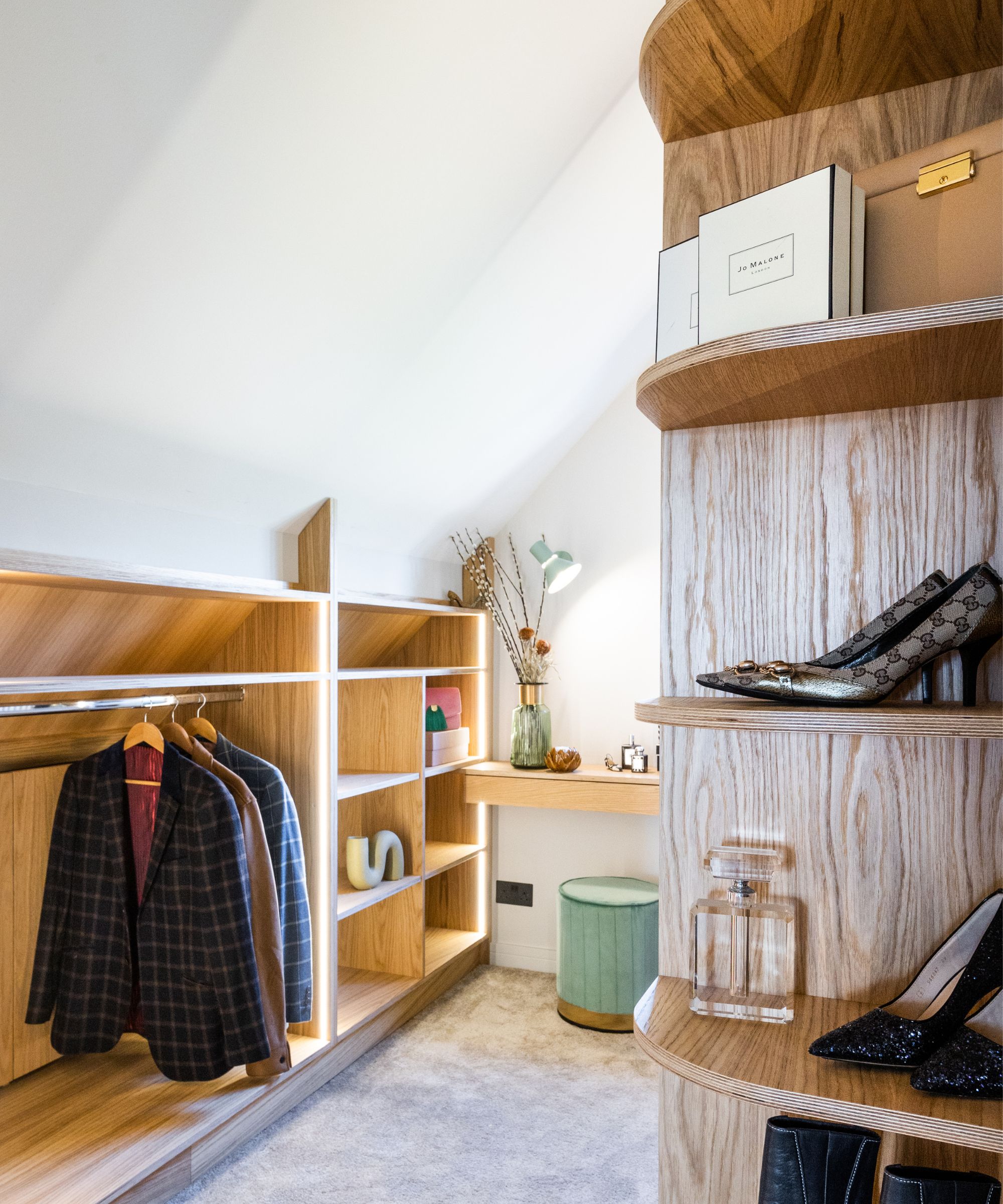A compact loft dressing room with sloped white ceilings and pale carpet. Light oak built-in shelving glows with soft strip lighting. Jackets hang neatly on a rail, while curved corner shelves display shoes and perfume bottles. A small vanity desk sits beneath.