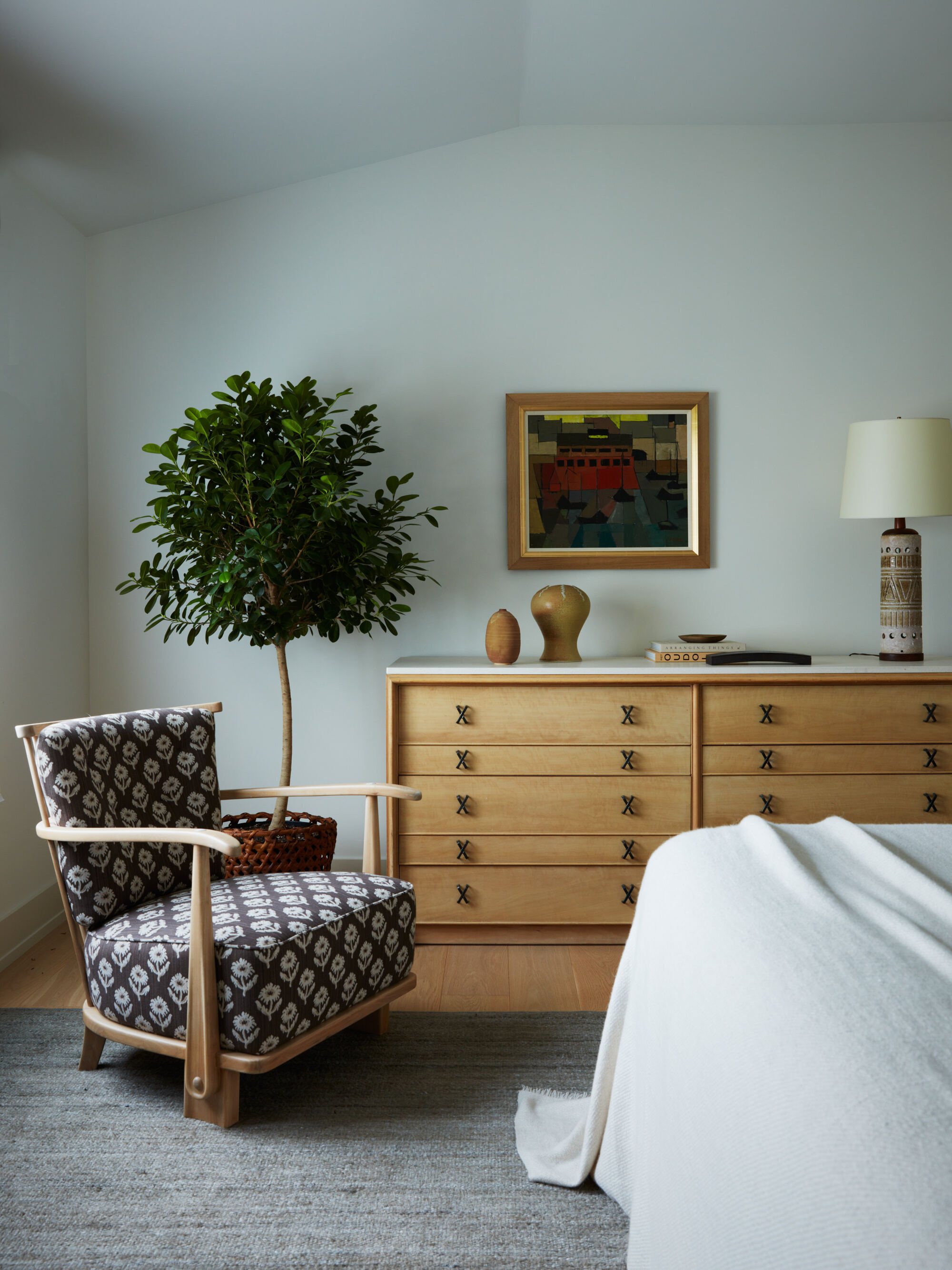 modern bedroom with light blue walls, timber console next to patterned armchair with potted tree, white bedding, and artwork on wall