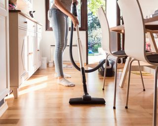 Female vacuuming hard kitchen floor between chairs and cabinets, with light coming in from back door behind her