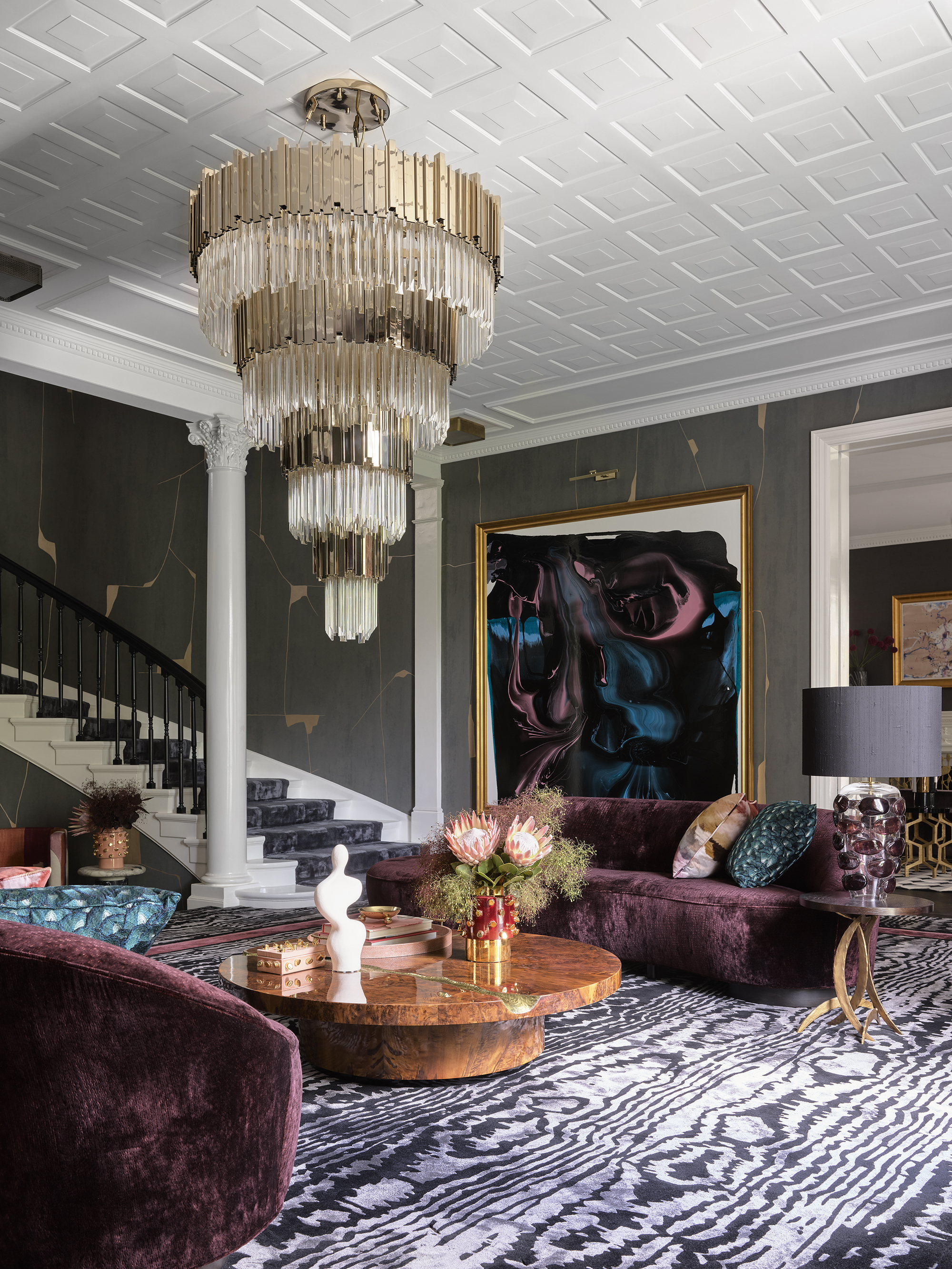 maximalist living room with velvet purple sofas, chandelier, patterned ceiling, animal print rug, green walls, curving staircase, burl wood coffee table styled with vases of flowers and sculptures, and a large artwork hanging on the wall