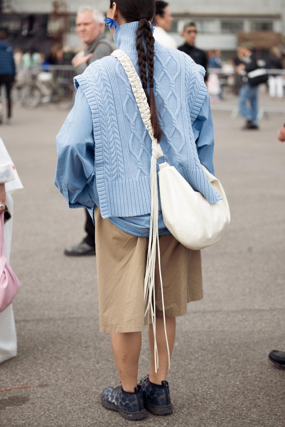 A guest wears beige shorts, blue shirt, blue knitted vest and white shoulder bag with tassels outside Cecilie Bahnsen during Copenhagen Fashion Week