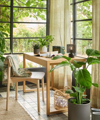 A cosy sunroom workspace with black-framed windows, a wooden desk, and lush greenery. Soft linen curtains and natural textures enhance the relaxed atmosphere.