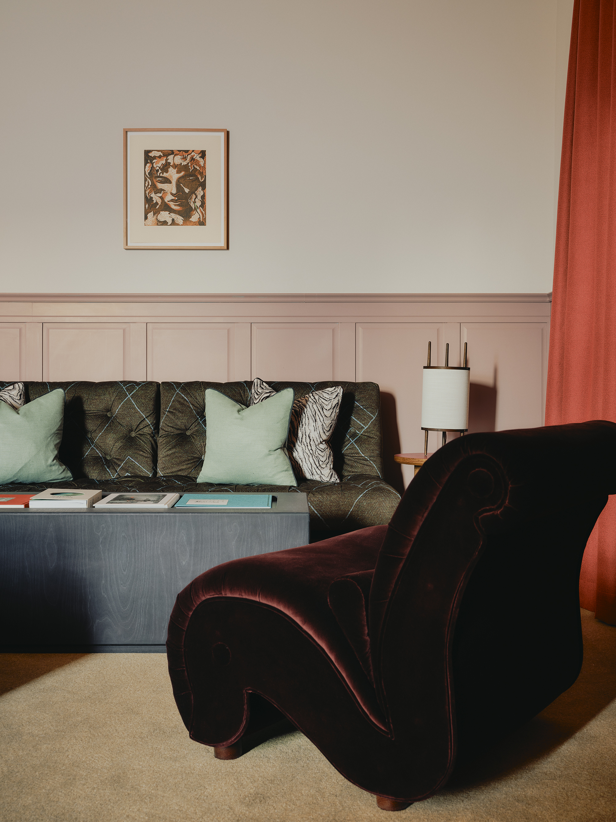 A hotel room decorated with velvety fabrics, across a two-person sofa and an armchair, pink-painted paneled walls, artworks, and statement lighting, all in colors such as mint green, red orange, and rose.