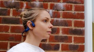 Aftershokz OpenRun headphones are one of the best headphones for cycling as they leave the ear open. This image shows a woman wearing a pair round her head, she is looking to the right and has a brick wall behind her